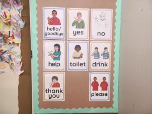 Classroom display showing illustrated Auslan signs for common words including hello, yes, no, help, toilet, drink, thank you and please.