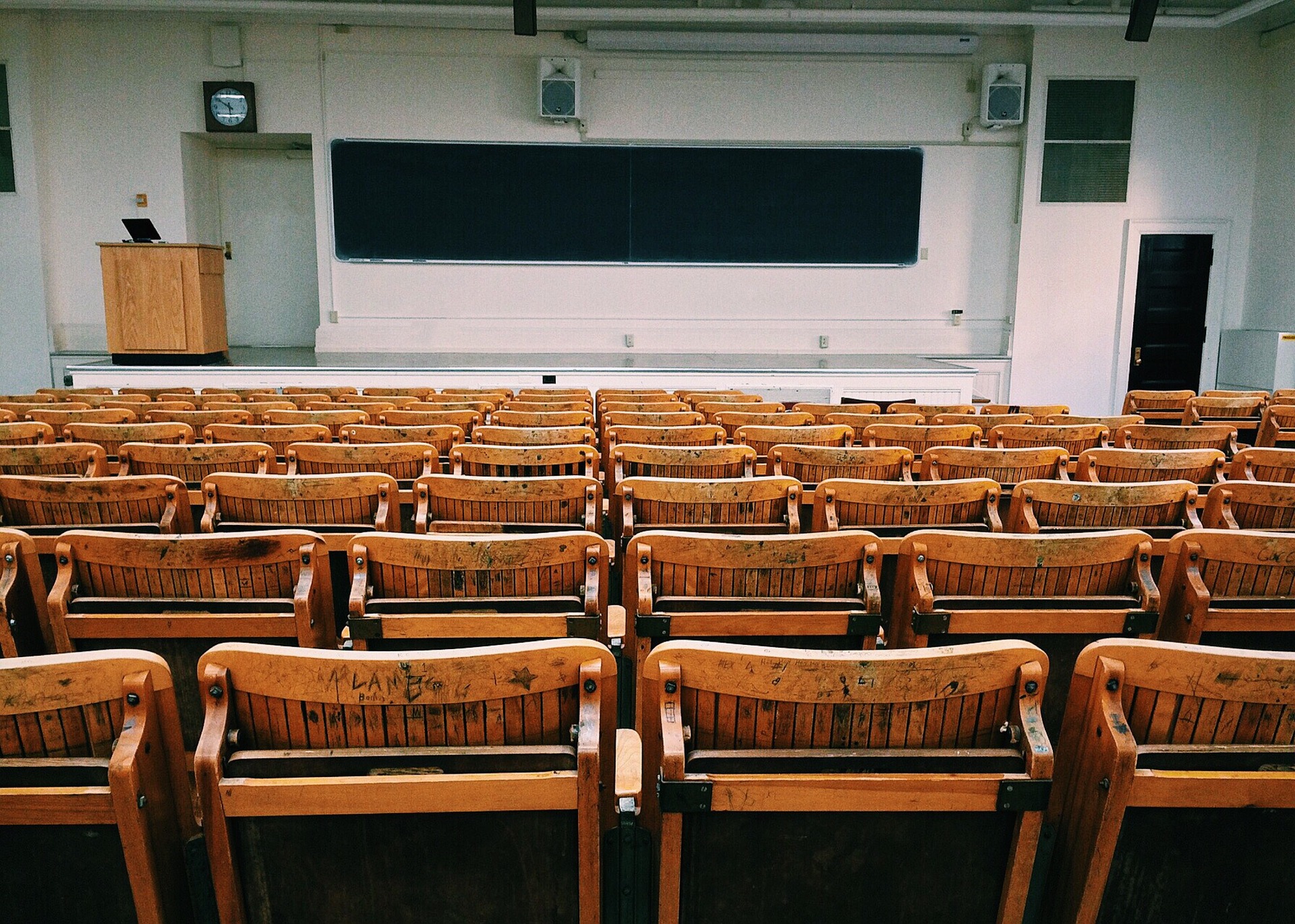 rows of wooden chairs facing a board and lectern in a lecture hall.